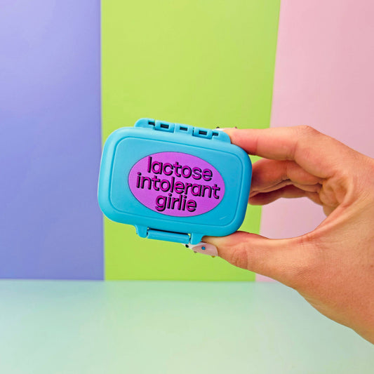 Blue pill organizer with 'lactose intolerant girls' text, strawberry milk container, and decorative items on a checkered background.