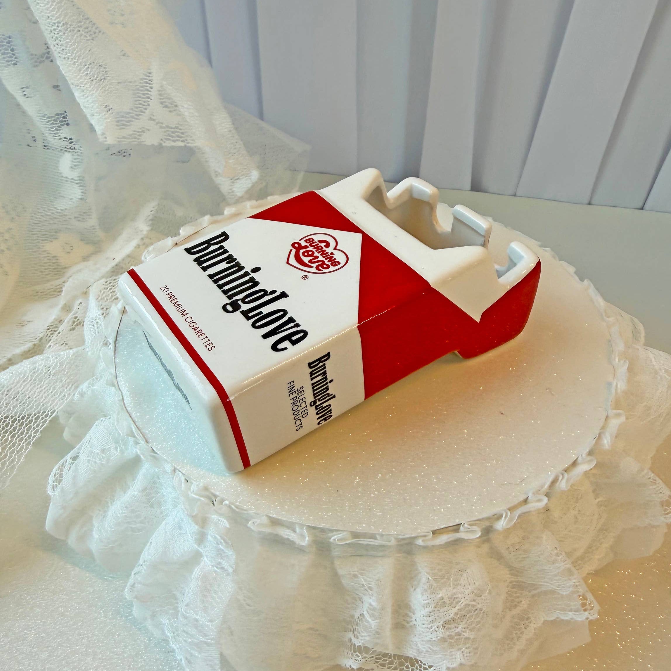 Kitsch faux cigarette box ash tray with a red label on a white lace surface