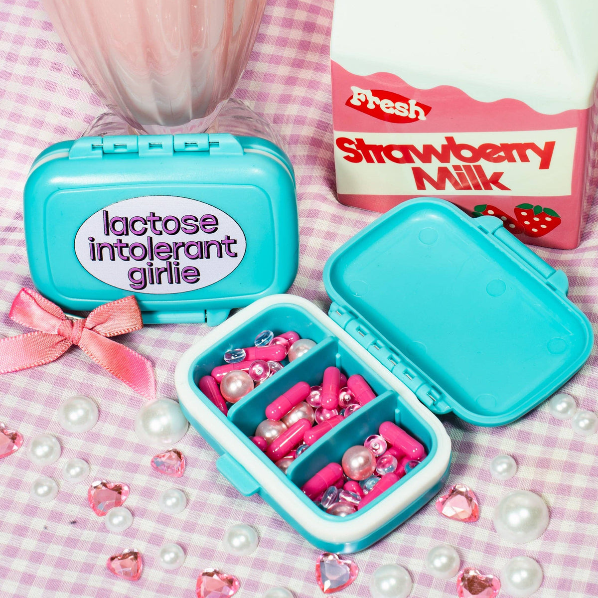 Blue pill organizer with 'lactose intolerant girls' text, strawberry milk container, and decorative items on a checkered background.