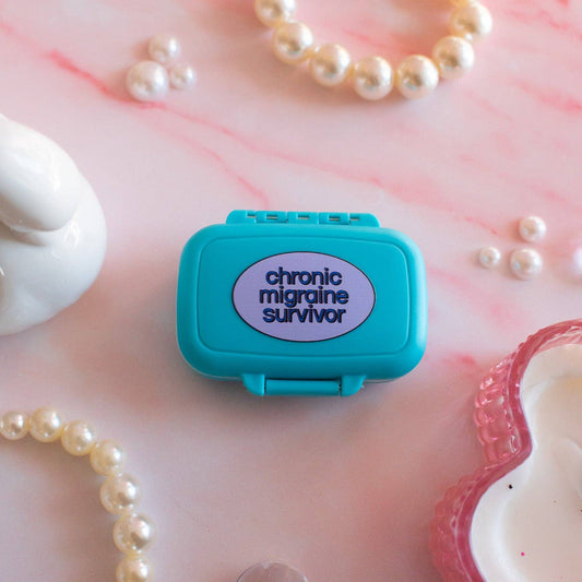 Teal pill box with 'chronic migraine survivor' text on a pink marble surface with pearls.