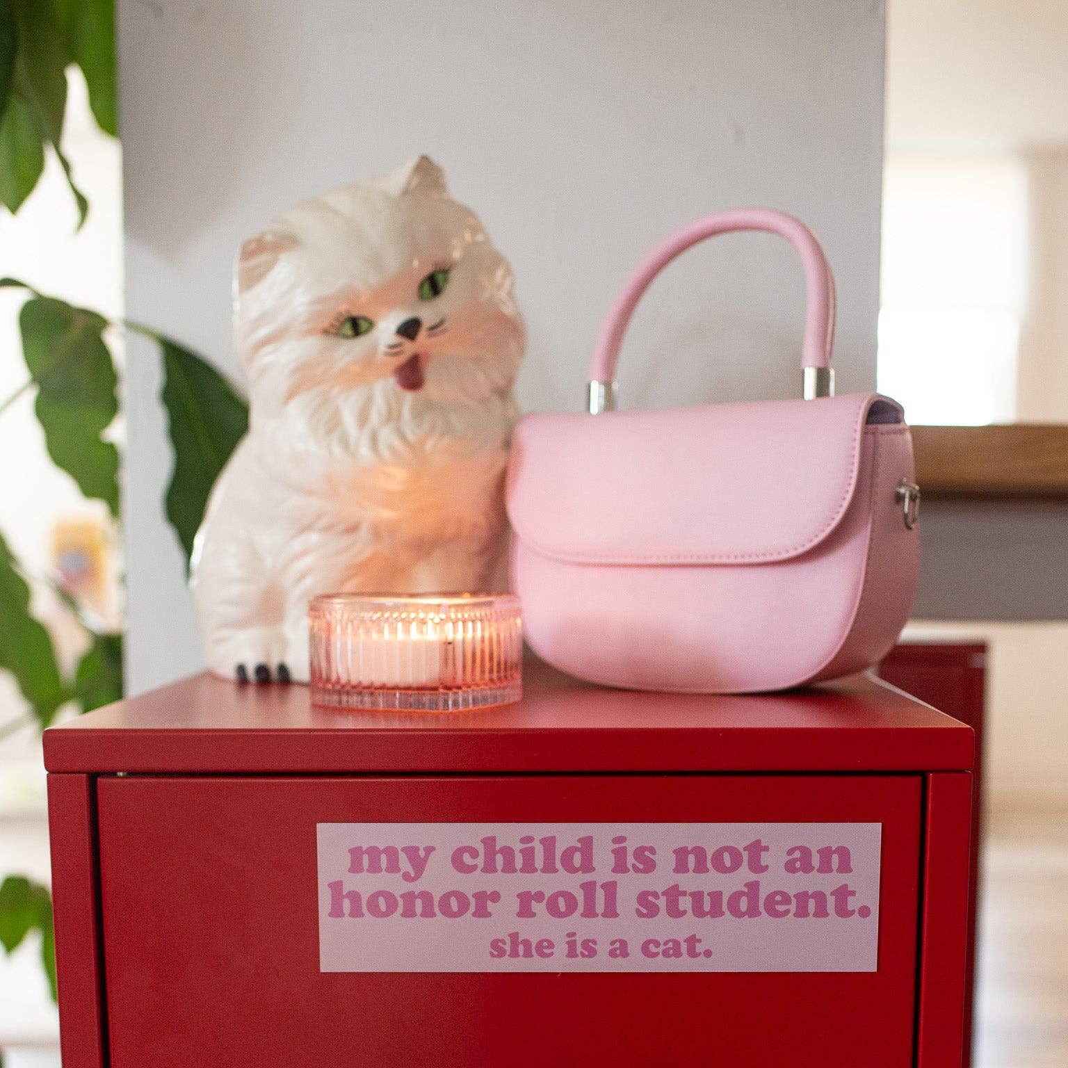 Pink cat-shaped lamp and handbag on a red surface with a humorous sign.