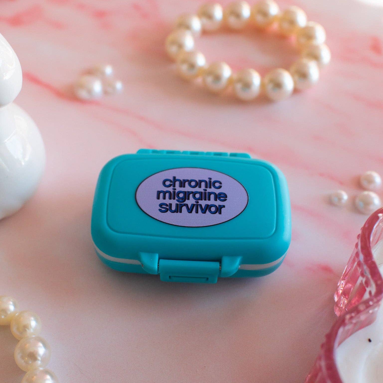 Small teal box with a purple label reading 'chronic migraine survivor' on a pink surface with pearls.