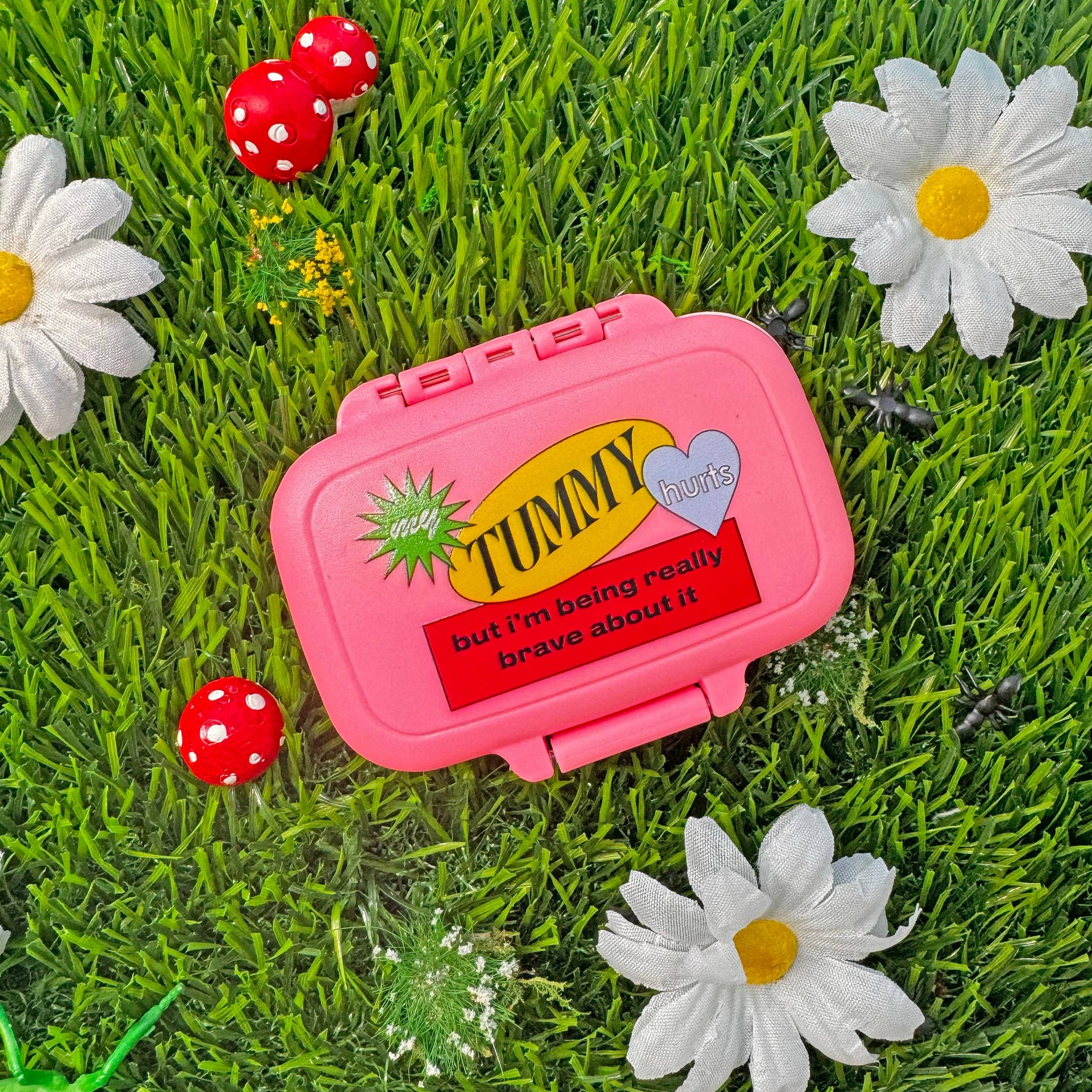 Pink 'Tummy' pill box on grass with flowers and ladybugs