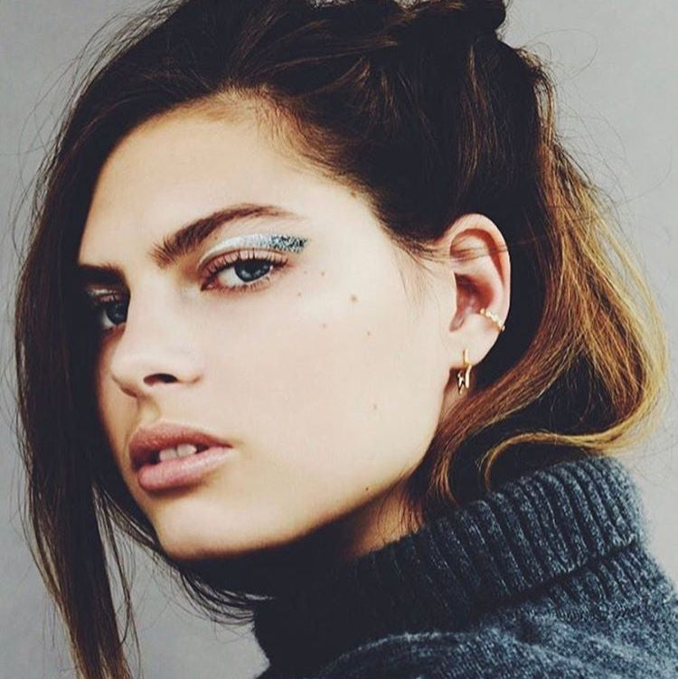Close-up of a person with stylish hair and makeup against a neutral background wearing tada and toy star hoop earrings
