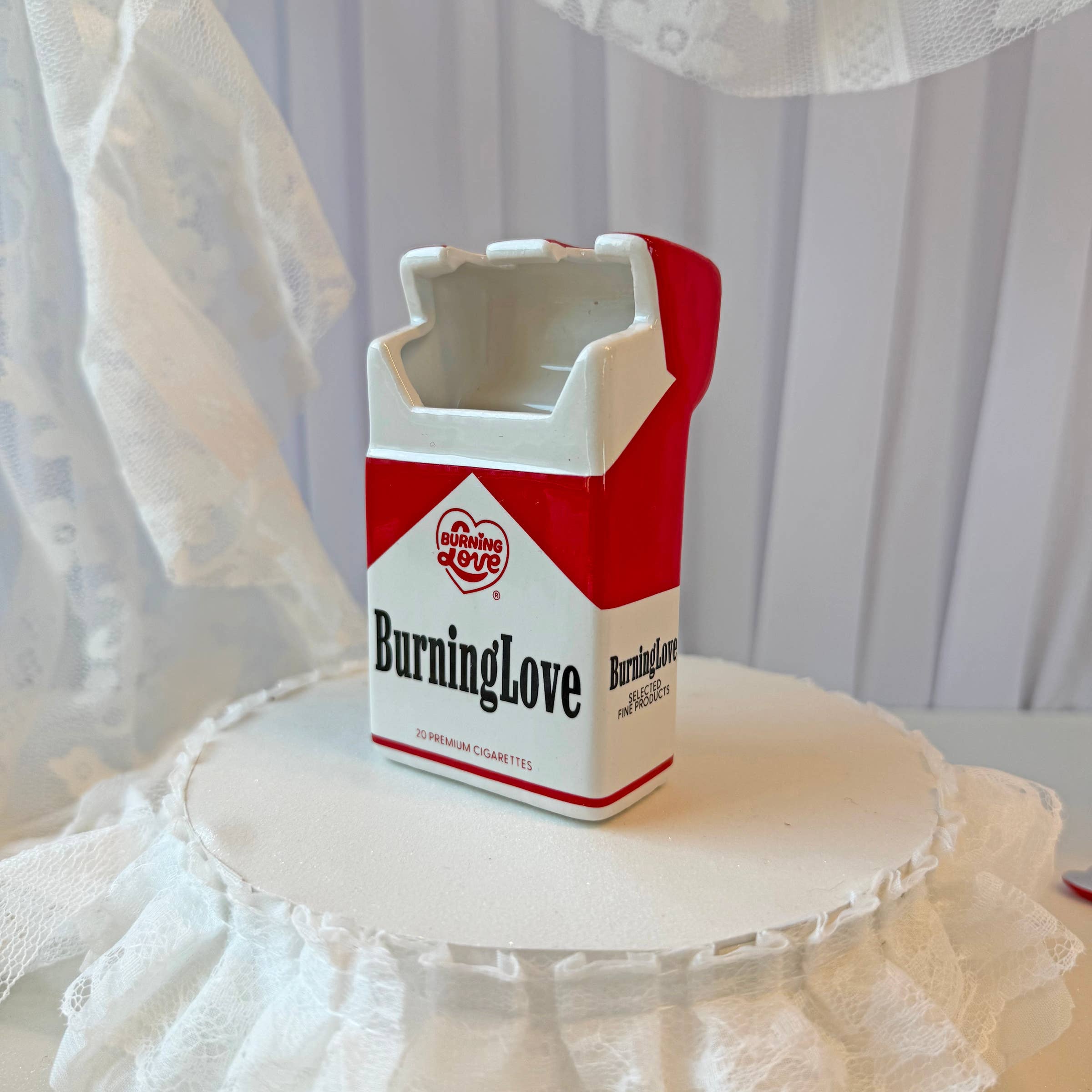 Cigarette carton-shaped ash tray / vase with 'Burning Love' text on a white surface with lace.