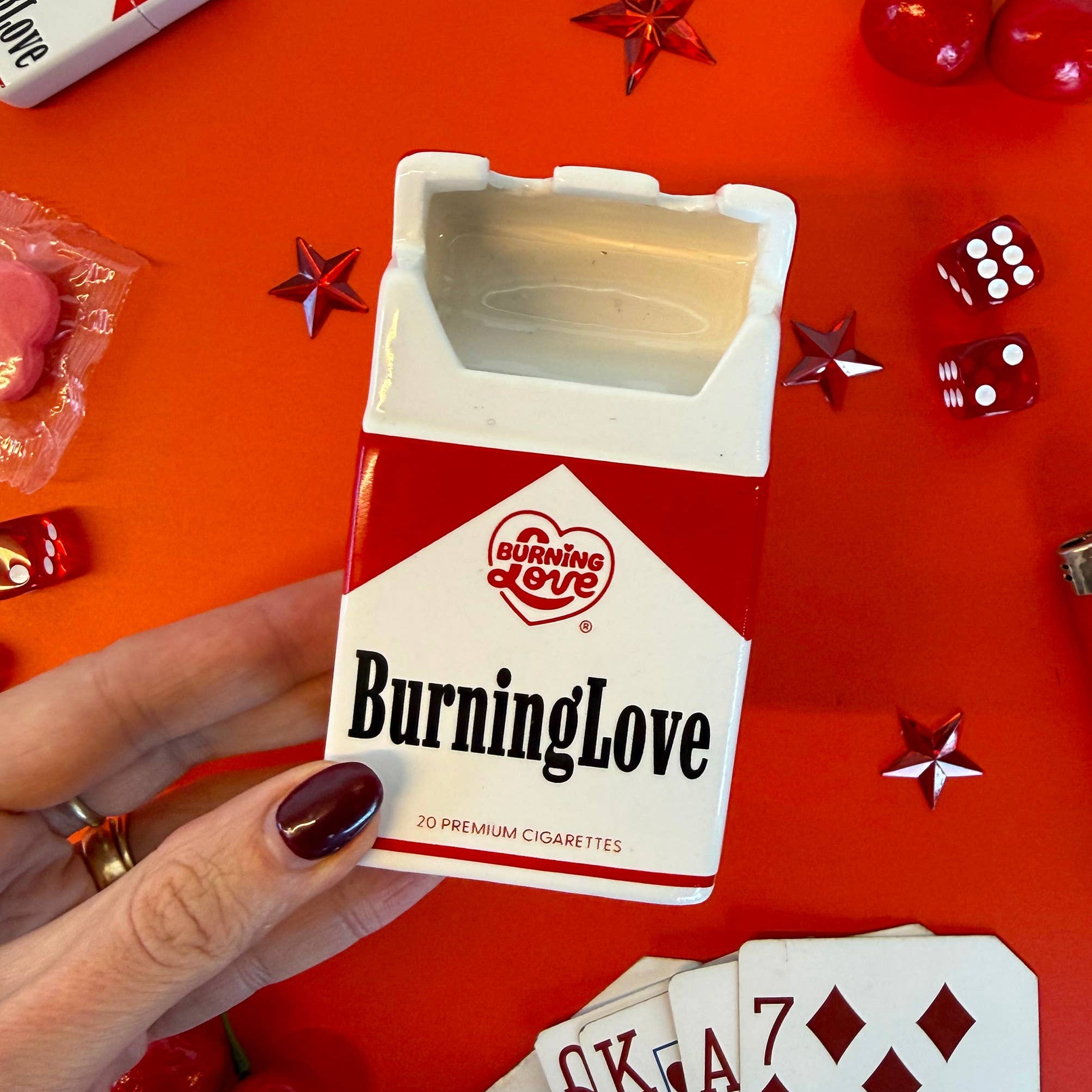 Hand holding a 'Burning Love' cigarette pack on a red background with playing cards and dice.