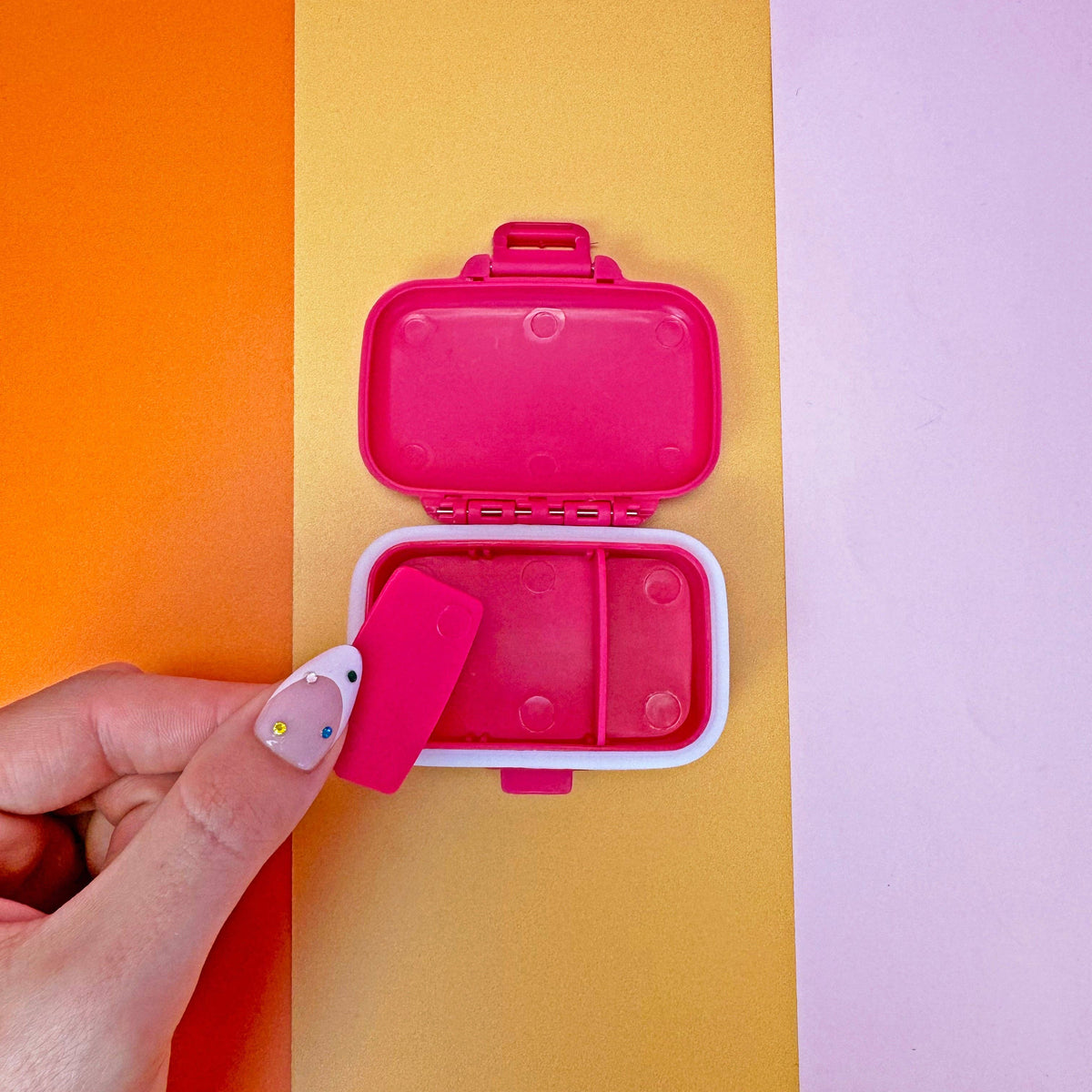 Pink pill box with an open lid on an orange, yellow, and purple background
