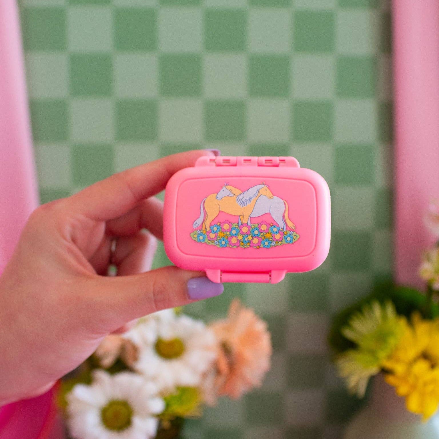 Hand holding a pink case with a kitsch horse design against a green checkered wall.