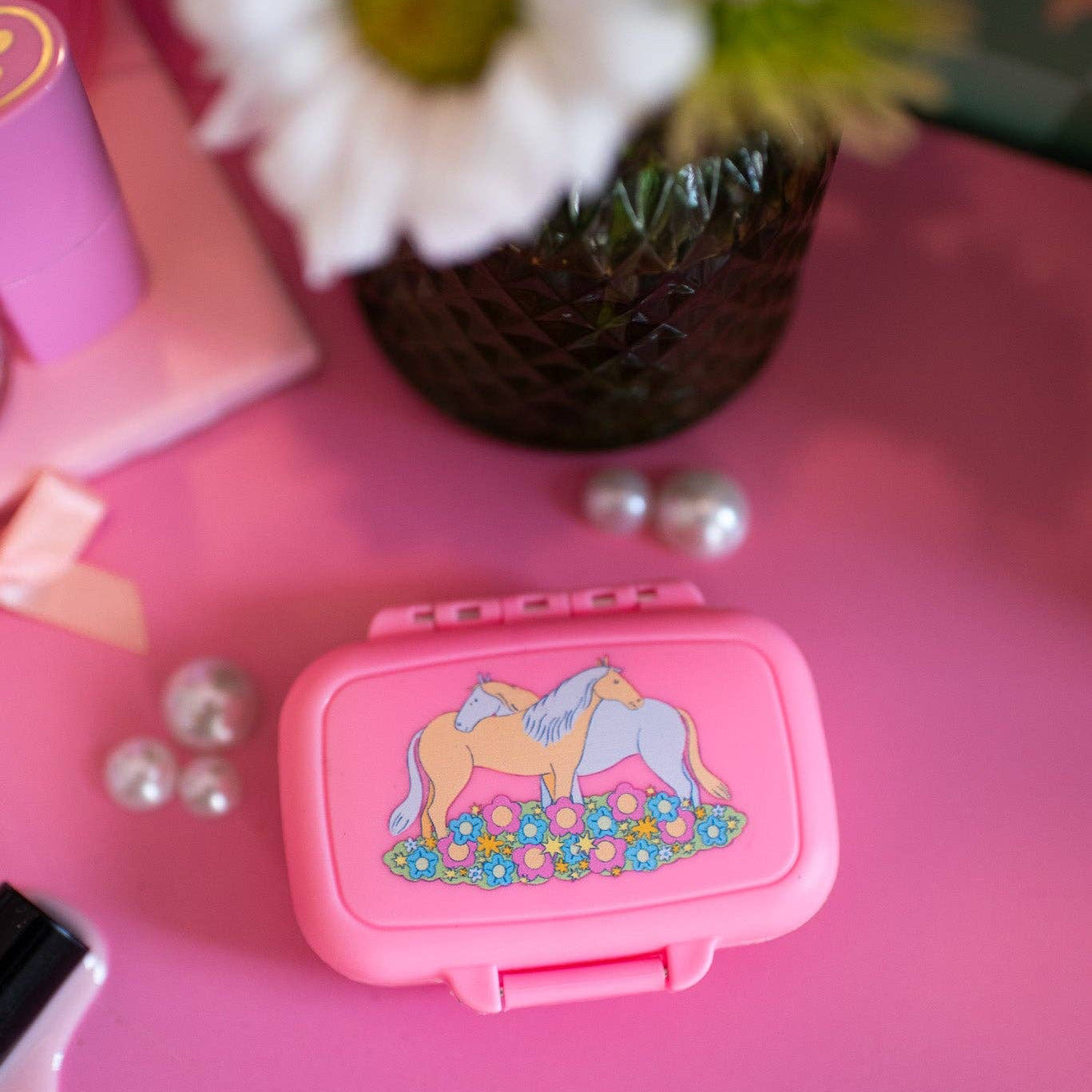 Pink pill box with horse design on a pink surface with flowers in the background