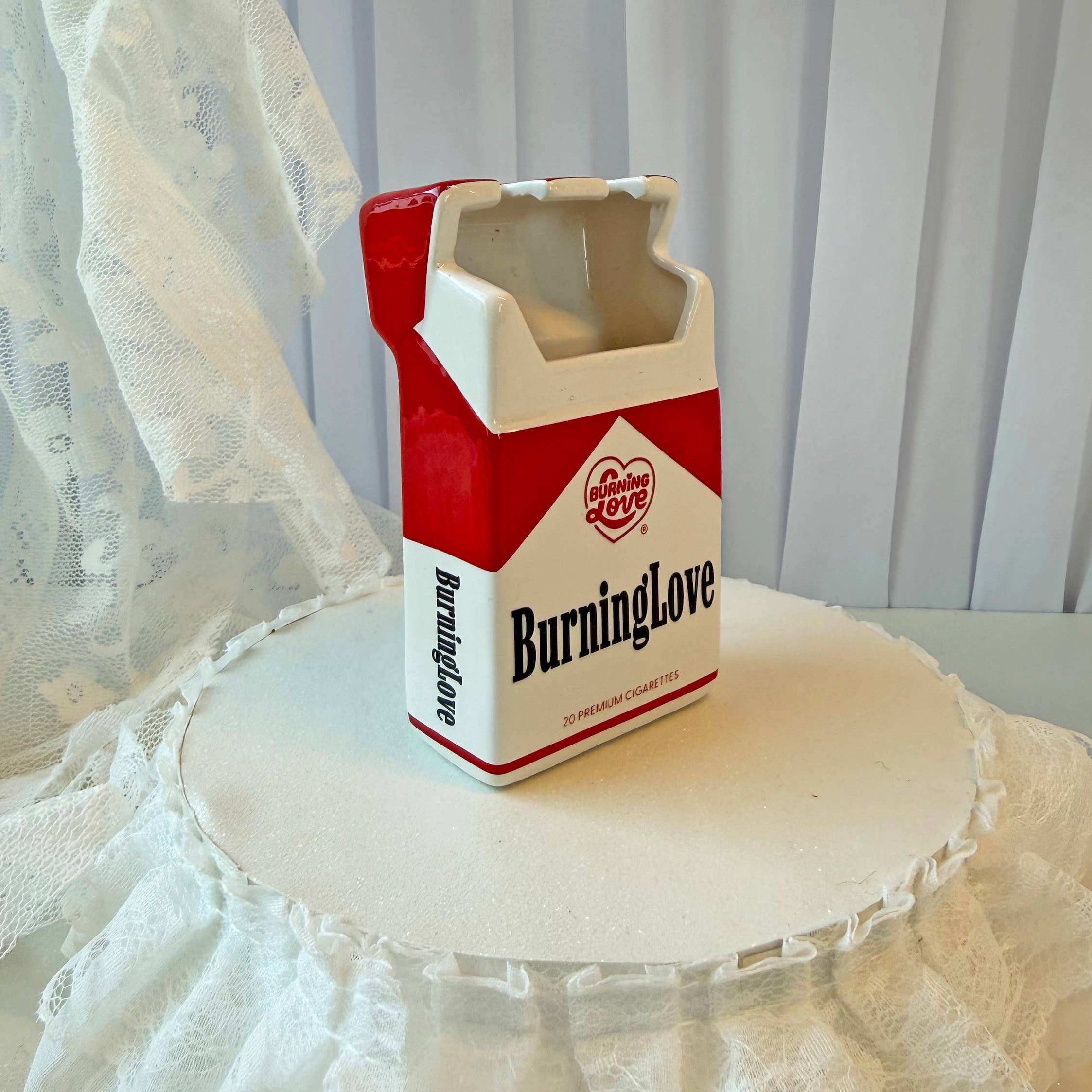 Cigarette pack-shaped container labeled 'Burning Love' on a white surface with lace curtains in the background.