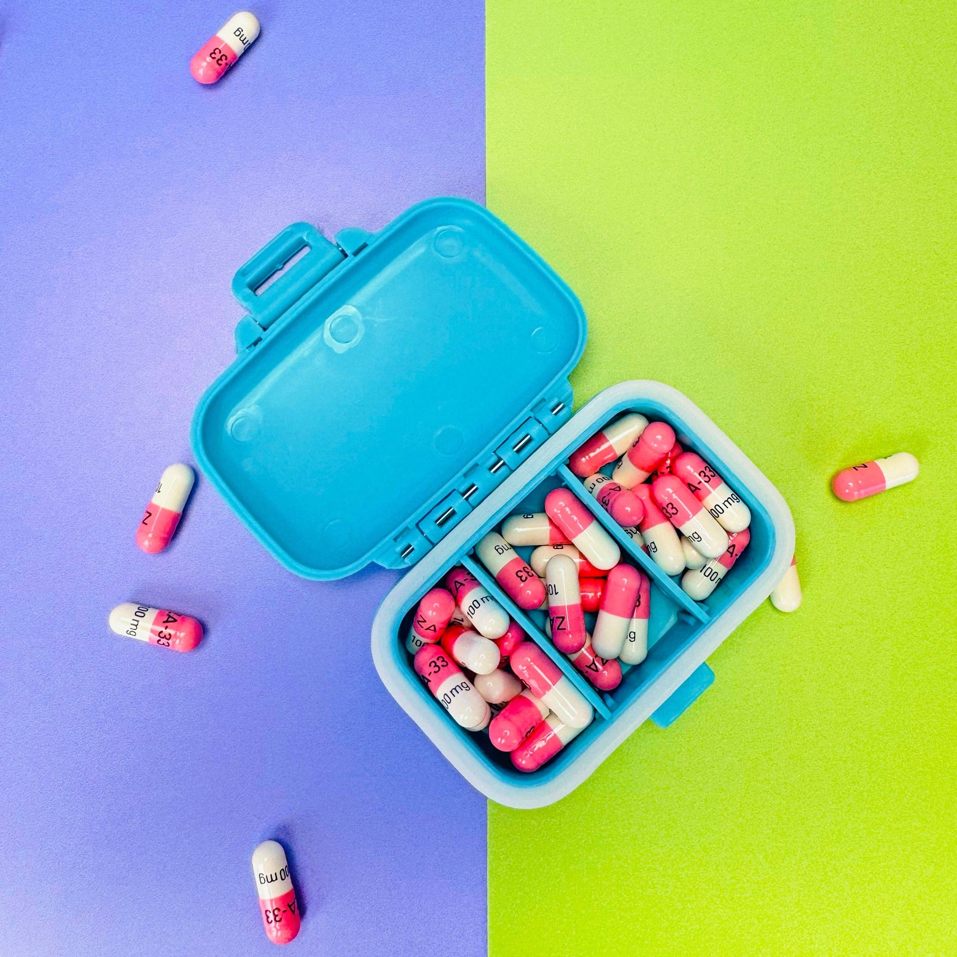 Blue pill organizer with pink and white capsules on a two-tone purple and green background