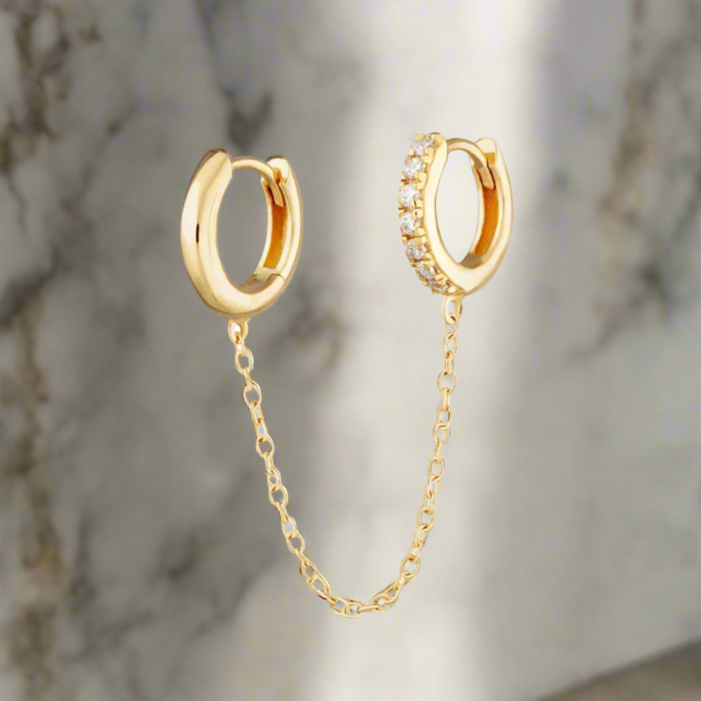 A pair of gold chain linked mismatched huggie hoop earrings with one hoop featuring a row of small crystals.
